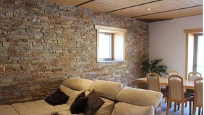 A living room with a stone wall, a beige sofa, and a dining area with wooden chairs and a table. - Grand Appartement Avec Loggia (Orchamps-Vennes)