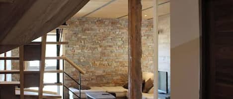 Un interior moderno con una escalera de madera que conduce a una zona superior tipo loft, una pared de piedra y una zona de estar acogedora con un televisor de pantalla plana.