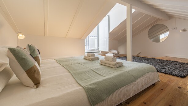 Een slaapkamer met een schuine plafond, een bed met een groene deken, een ronde spiegel en een houten vloer.