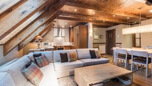 Ein gemütliches Wohnzimmer im Loft-Stil mit Holzbalkendecke, einer Sofagarnitur, einem Esstisch mit Stühlen und einer Küche im Hintergrund.