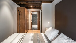 Un dormitorio con techo de madera, una cama con ropa de cama estampada, una mesita de noche con lámpara, y una puerta que da a un baño con ducha.