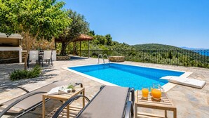 Una piscina con sillones de descanso y una mesa con bebidas, rodeada de pavimento de piedra y follaje.