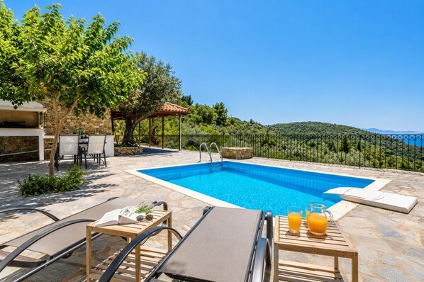 A swimming pool with lounge chairs and a table with drinks, surrounded by stone flooring and greenery. - Oak Tree Cottage - One Bedroom Villa, Sleeps 2 (Sporades)