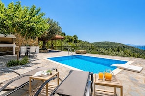 Una piscina con sillones de descanso y una mesa con bebidas, rodeada de pavimento de piedra y follaje.