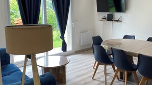 Moderni huone, jossa on pyöreä puinen pöytä, siniset tuolit, suuri lattiavalaisin, seinälle asennettu taulutelevisio ja ikkuna, jossa on verhot.