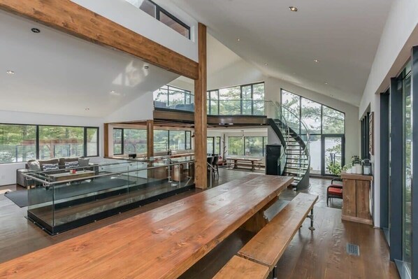 A spacious interior with a long wooden table, a glass railing, and a staircase leading to an upper level.