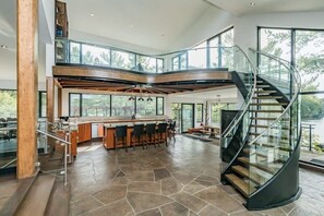 A modern kitchen with a central island, a curved staircase, and large windows overlooking a natural landscape.