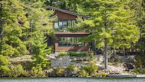 A modern house with a stone foundation, large windows, and a dock with chairs. - Cherry Island Muskokas Premiere Private Island (Utterson)