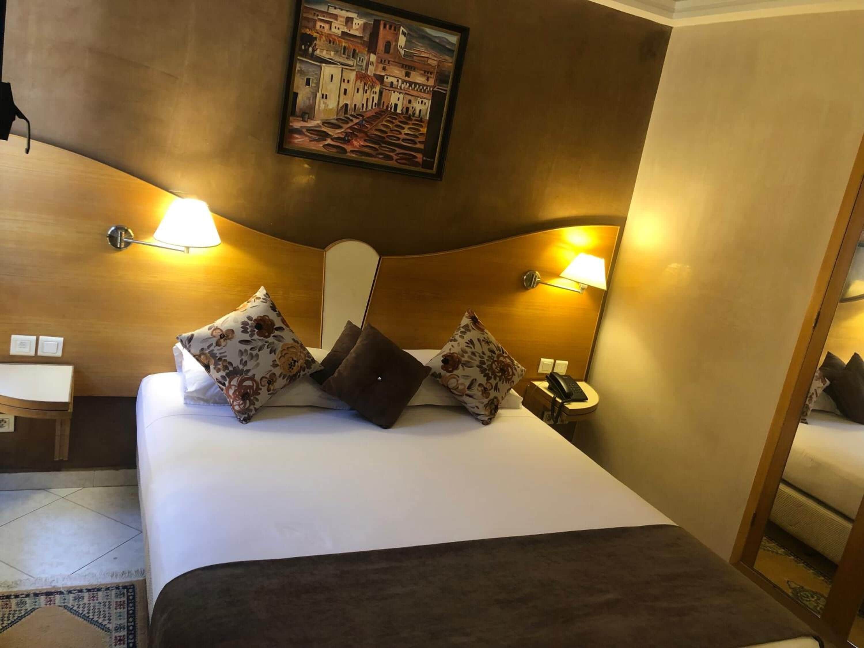 A hotel room with a large bed, two bedside lamps, a painting on the wall, and a small table with a chair.