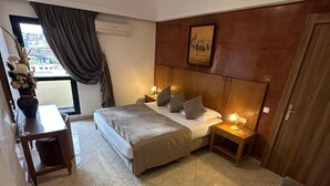 A hotel room with a bed, bedside tables, a desk, a chair, a window with curtains, and a painting on the wall. - Mounia (Fes)