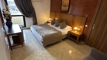A hotel room with a bed, bedside tables, a desk, a chair, a window with curtains, and a painting on the wall.