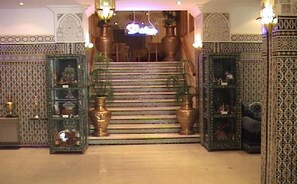 Una escalera con barandillas ornamentales y paredes con diseños, que conduce a una habitación con candelabro.