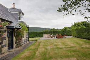 A stone house with a black door, a wooden hot tub, and a lawn with a patio table and chairs. - Traditional Highland Family Cottage with Hot Tub on Mayen Estate, Banffshire (Huntly)
