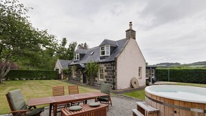 Une maison avec une façade en pierre, une terrasse en bois, un jacuzzi et une pelouse.