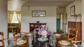 Une salle à manger avec une longue table dressée pour six personnes, entourée de chaises en bois, un vase de fleurs et une carte encadrée accrochée au mur.