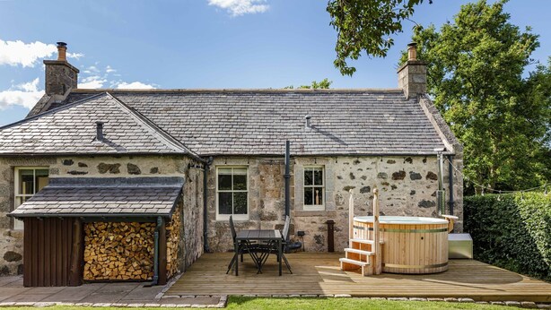 A stone house with a tiled roof, a wooden deck, a hot tub, and a stack of firewood. - Chic Traditional Highland Estate Cottage with Valley View and Hot Tub (Rothiemay)