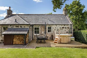 A stone house with a tiled roof, a wooden deck, a hot tub, and a stack of firewood.