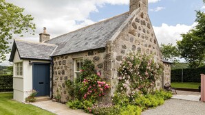 A stone cottage with a blue door, surrounded by a well-maintained garden with pink flowers. - Chic Traditional Highland Estate Cottage with Valley View and Hot Tub (Rothiemay)