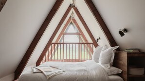 Un dormitorio con techo inclinado, estructura de cama de madera, mesita de noche con lámpara y vistas al exterior a través de una ventana.