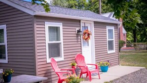 Une petite maison avec un porche, deux chaises Adirondack rouges, une table et des plantes en pot.