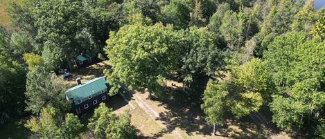 An aerial view of a cabin with a green roof, surrounded by dense forest and a lake.