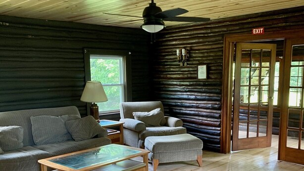 Ein Interieur einer Blockhütte mit Holzbalkendecke, Deckenventilator, einer Couch, einem Couchtisch, einer Lampe und einer Tür.