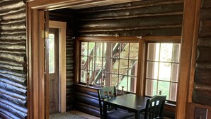 Ein rustikales Holzhaus-Interieur mit einem Esstisch, Stühlen, einem grünen Sessel und einem Hinweisschild für den Ausgang.