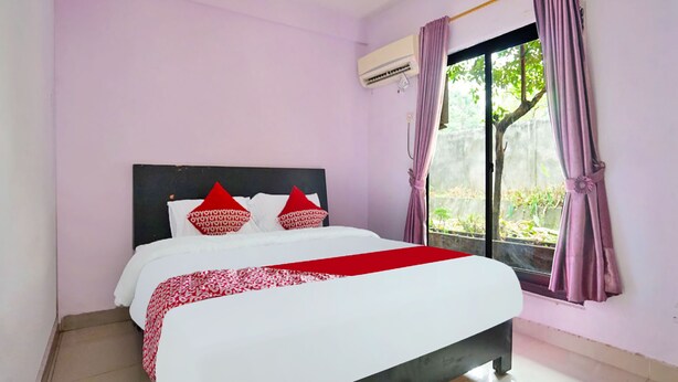 Een net opgemaakt bed met een wit en rode sprei, twee rode kussens en een zwart hoofdbord. Er is een raam met paarse gordijnen en een airconditioningunit aan de muur bevestigd.