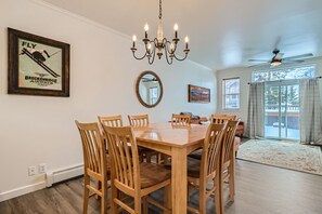 Uma sala de jantar com mesa e cadeiras de madeira, um lustre, um cartaz emoldurado, um espelho e vista para o exterior através de uma porta de vidro deslizante.