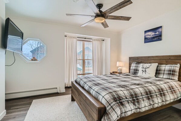 Quarto com cama de madeira, ventilador de teto, televisão montada e janela com cortinas.