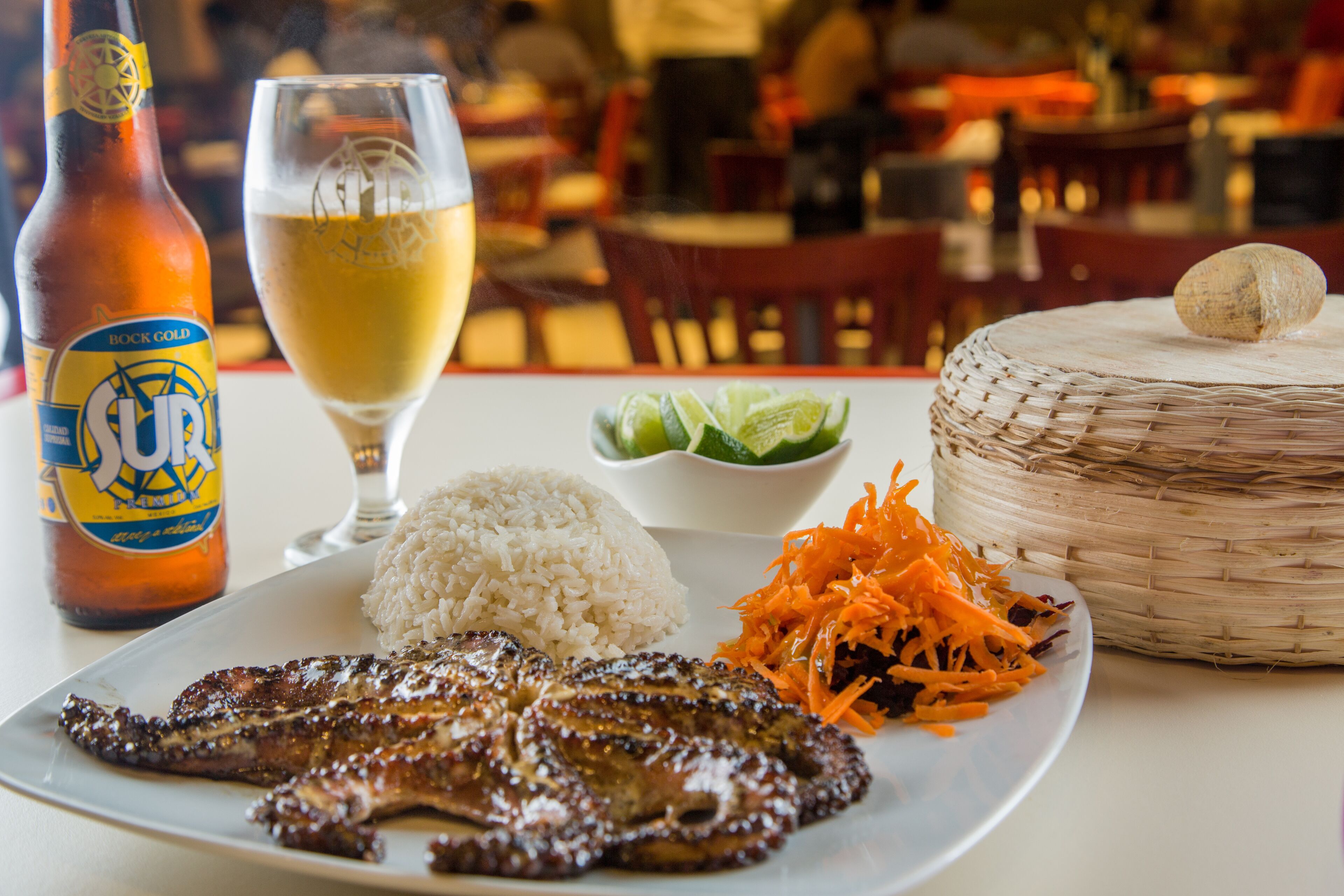 A plate with grilled meat, rice, and shredded carrots, a glass of beer, and a bottle of beer on a table in a restaurant.