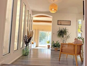 A spacious hallway with wooden flooring, large windows, and a rattan chair.