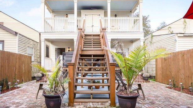 A two-story house with a wooden staircase leading to a front door, surrounded by a brick patio and potted plants.