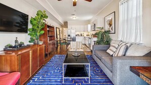 A modern living room with a sofa, coffee table, and a TV. There is a ceiling fan, a kitchen in the background, and a blue patterned rug.
