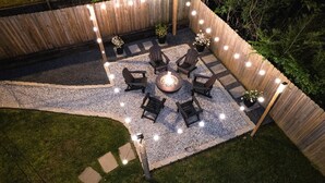 An outdoor patio area with a fire pit surrounded by Adirondack chairs, a wooden fence, and string lights.