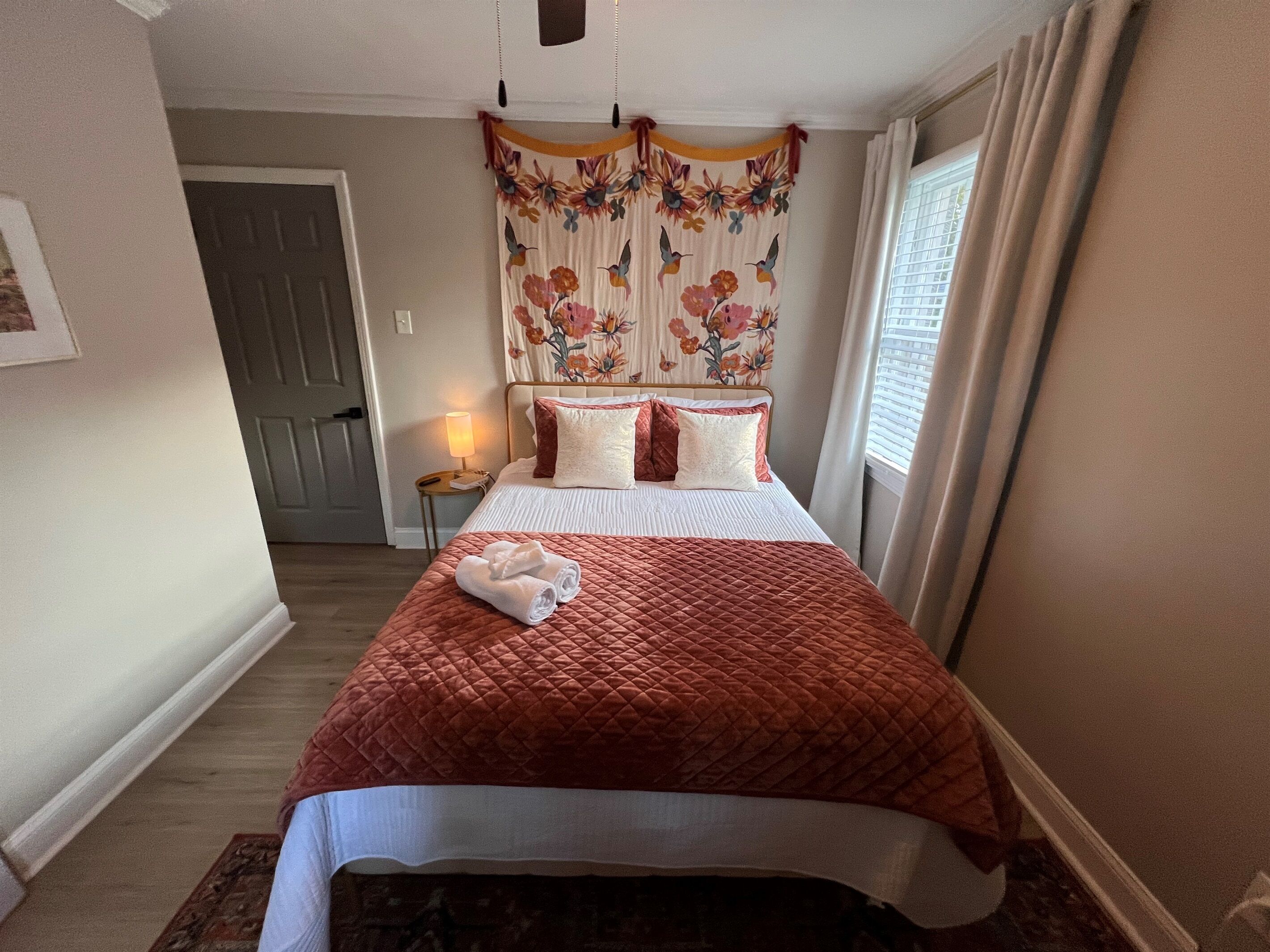A neatly made bed with a red quilt, a bedside lamp, and a floral curtain.