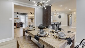 A dining area with a long wooden table set for a meal, a modern chandelier, and a view into another room with a kitchen and seating area.