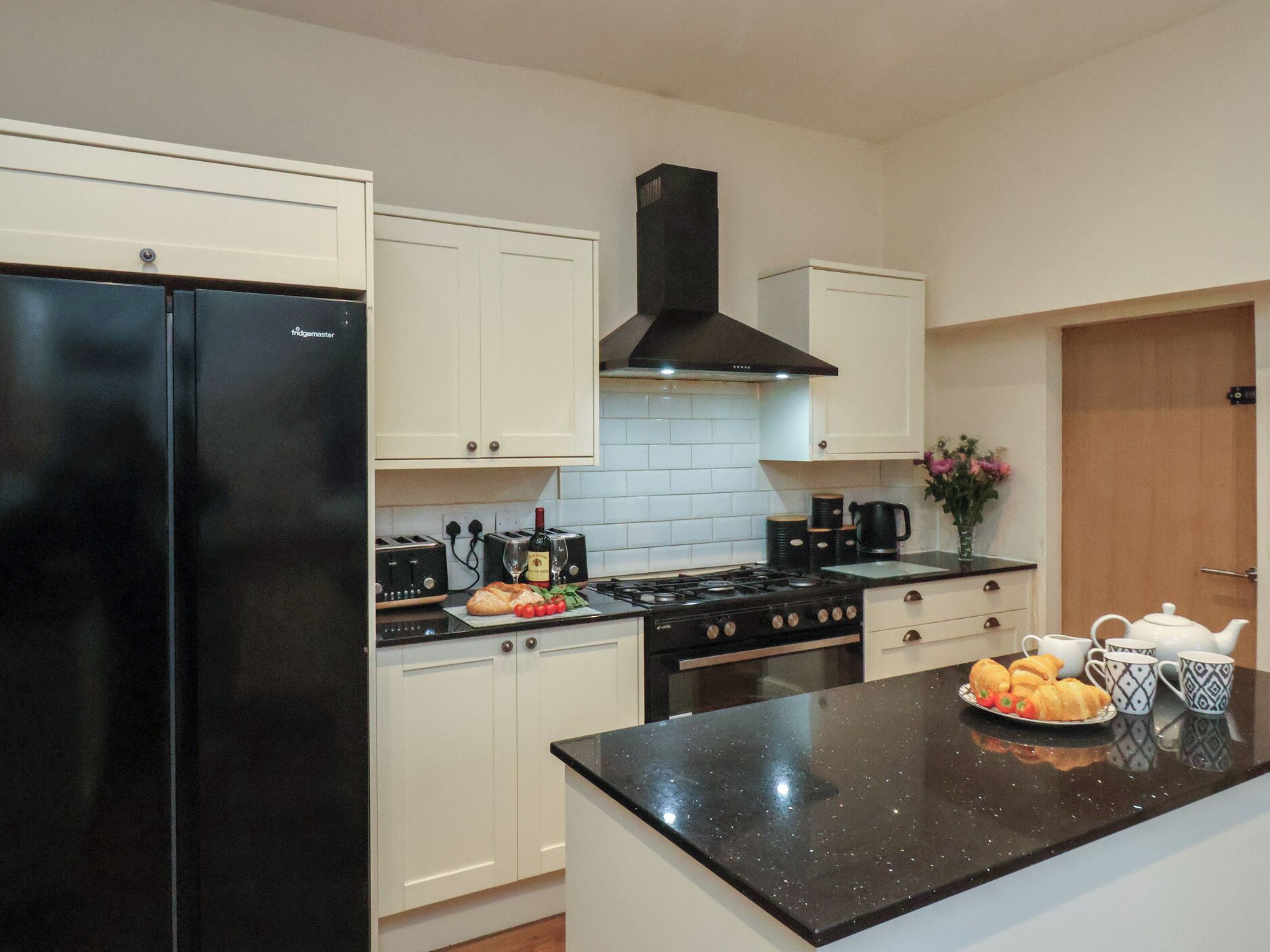 Una cocina moderna con armarios blancos, un refrigerador negro, una estufa y una encimera con fruteros y una tetera.