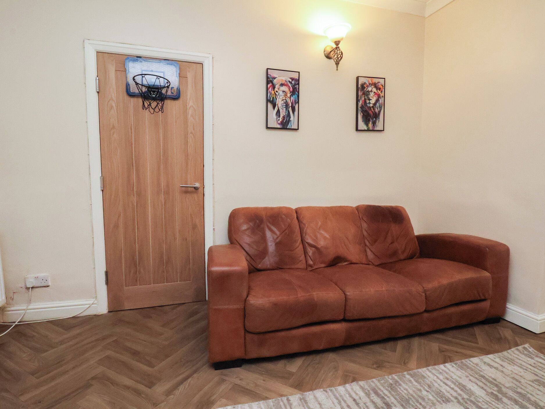 Une pièce avec un canapé en cuir marron, une porte en bois, un panier de basket fixé au mur et deux œuvres d’art encadrées.