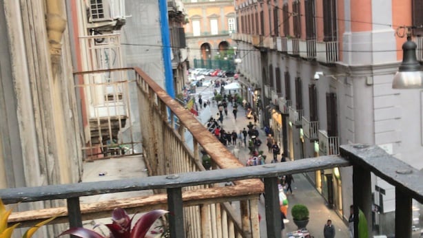 A narrow street with buildings on either side, a pedestrian walkway, and potted plants in the foreground. - Base Napoli Plebiscito (Naples)