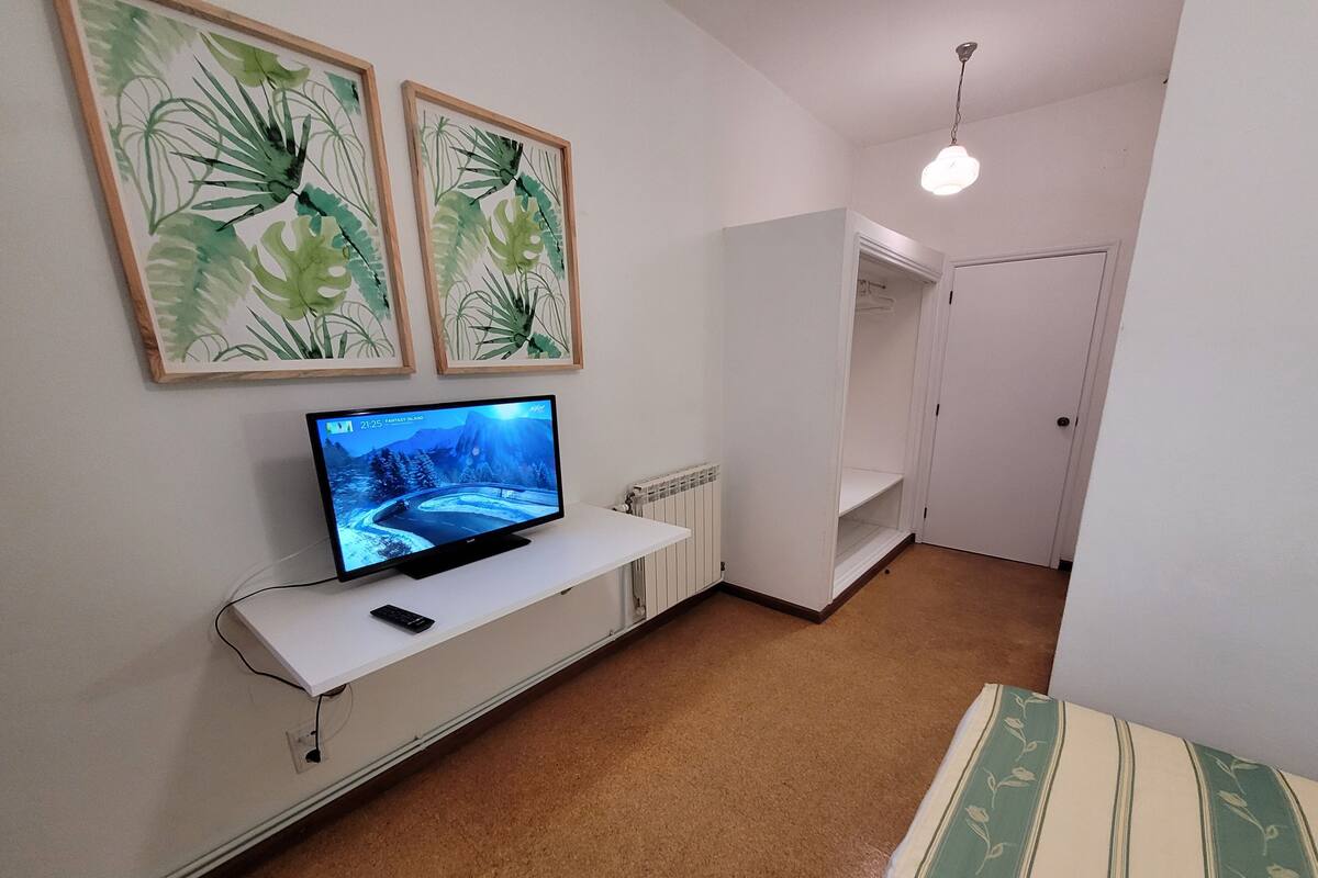 Een kamer met een televisie op een witte standaard, een afstandsbediening, en twee wanddecoraties met bladmotieven.