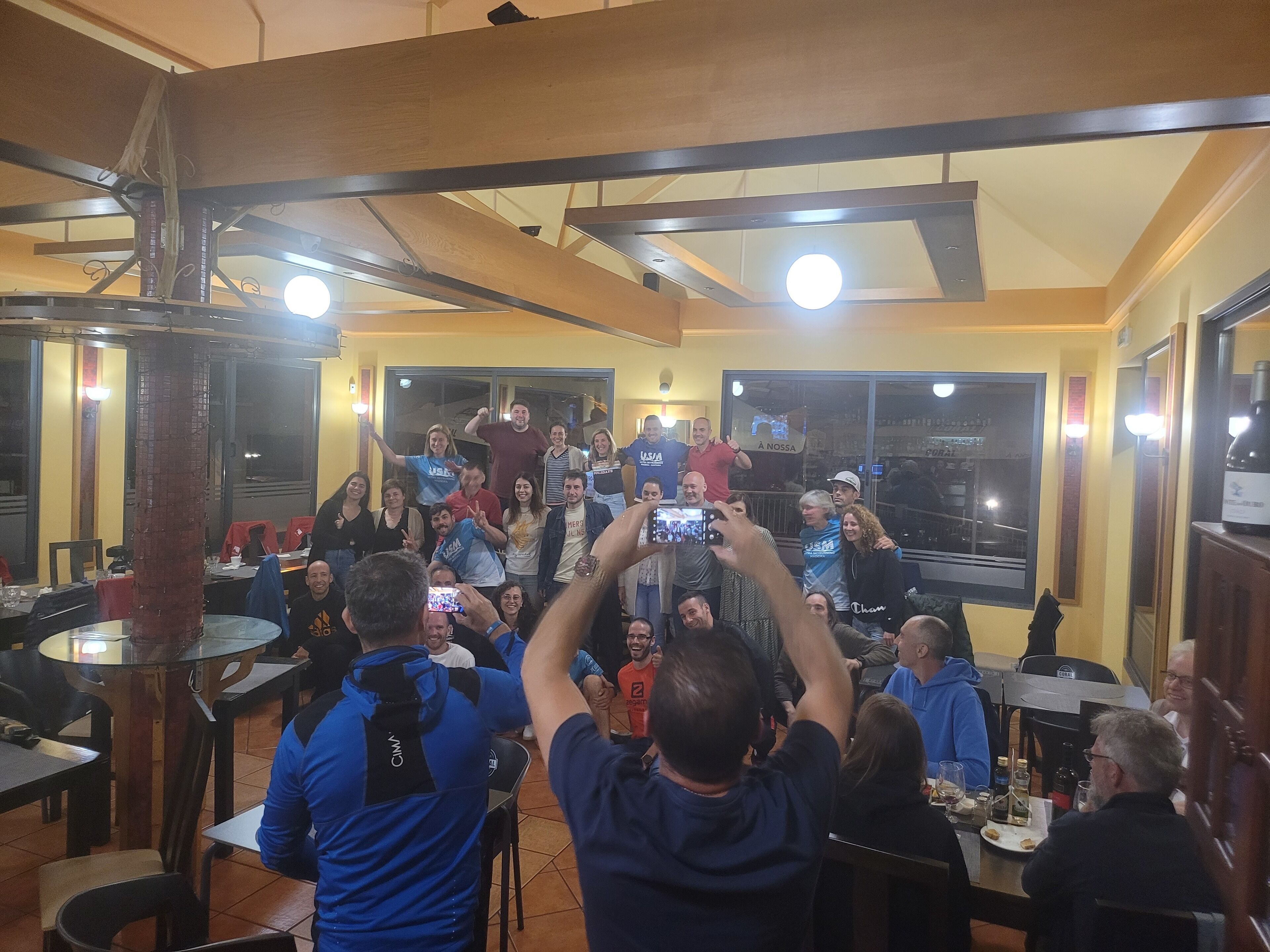 A restaurant with a high ceiling, exposed beams, and large windows. Tables are arranged around the room, and people are gathered in the center, some taking photos.