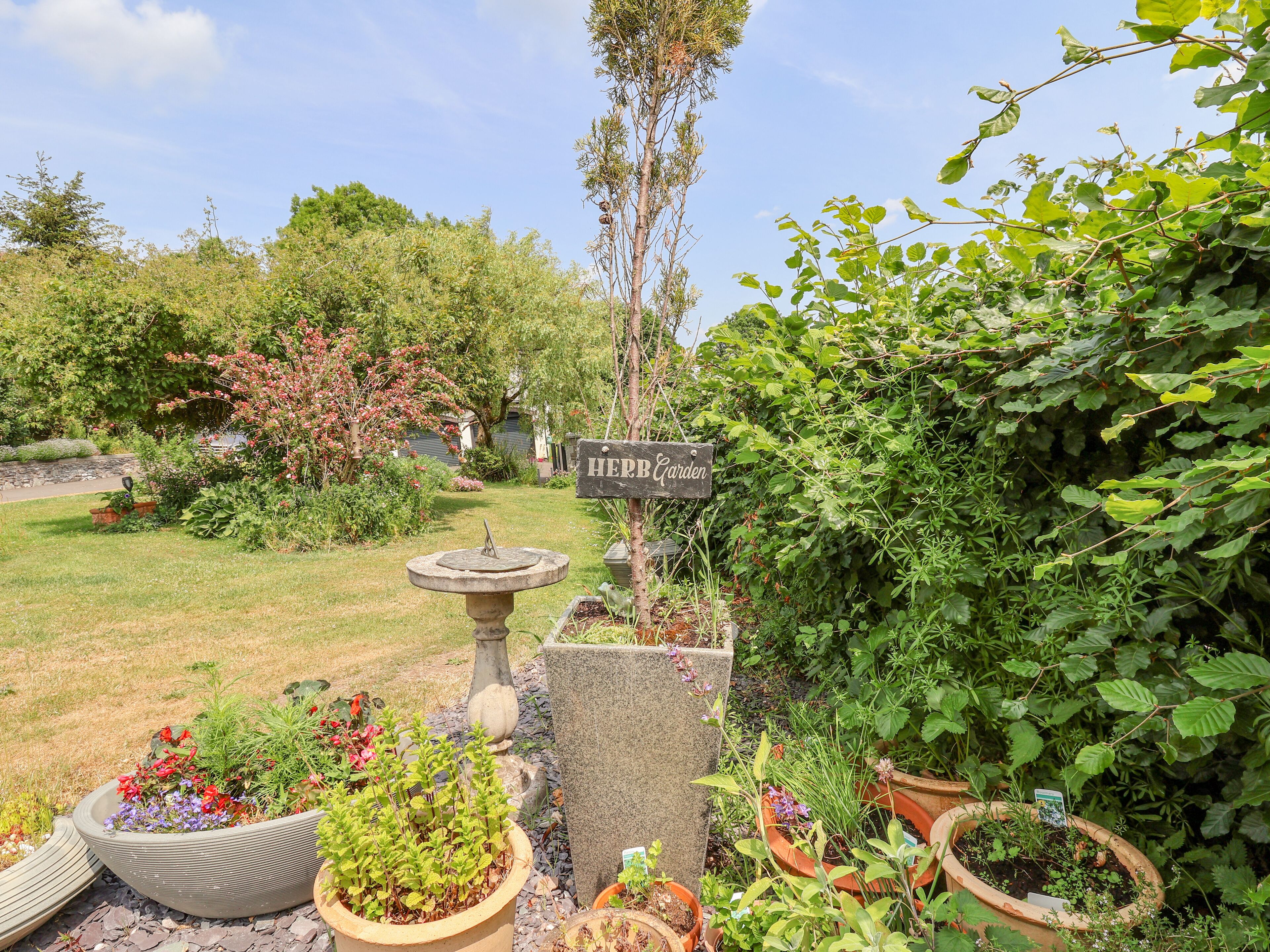 Ein Garten mit Sonnenuhr, einem Schild mit der Aufschrift 'HERB Garden' und verschiedenen Blumentöpfen.