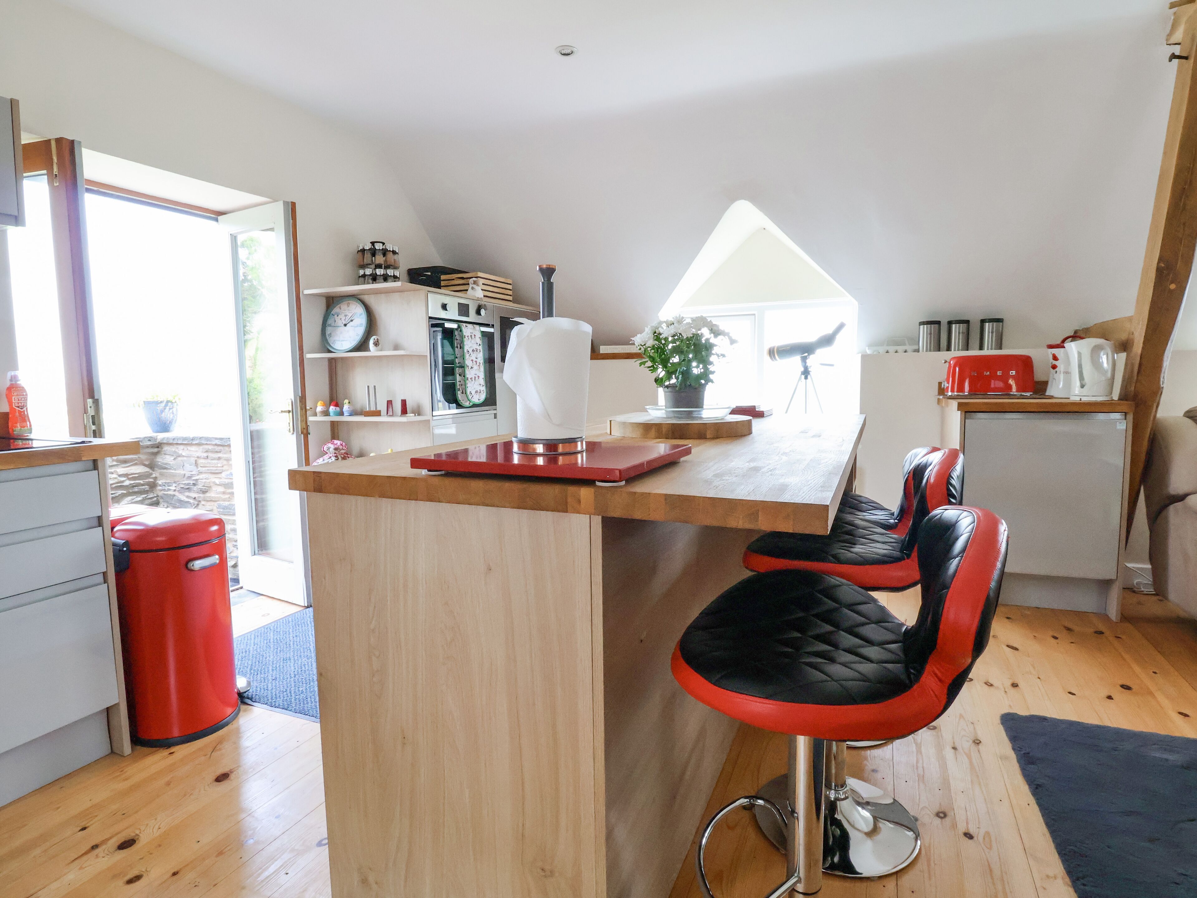 Une cuisine avec un îlot central en bois, des tabourets de bar rouges, une poubelle rouge et une vue sur l’extérieur.