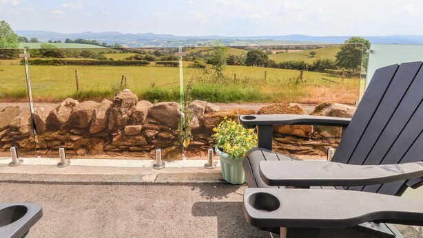 Un patio con un muro de piedra, una planta en maceta y dos sillones de descanso mirando una vista panorámica de un campo verde y montañas lejanas.