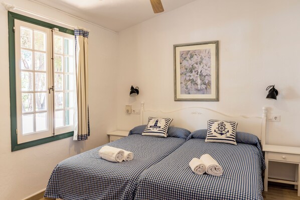 A bedroom with two beds, checkered bedding, a framed floral artwork, and a window with curtains. - Las Brisas I and II Apartments (Ciutadella de Menorca)