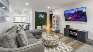 Un salon moderne avec un canapé d’angle gris, un pouf à motifs, un téléviseur à écran plat et une bibliothèque.