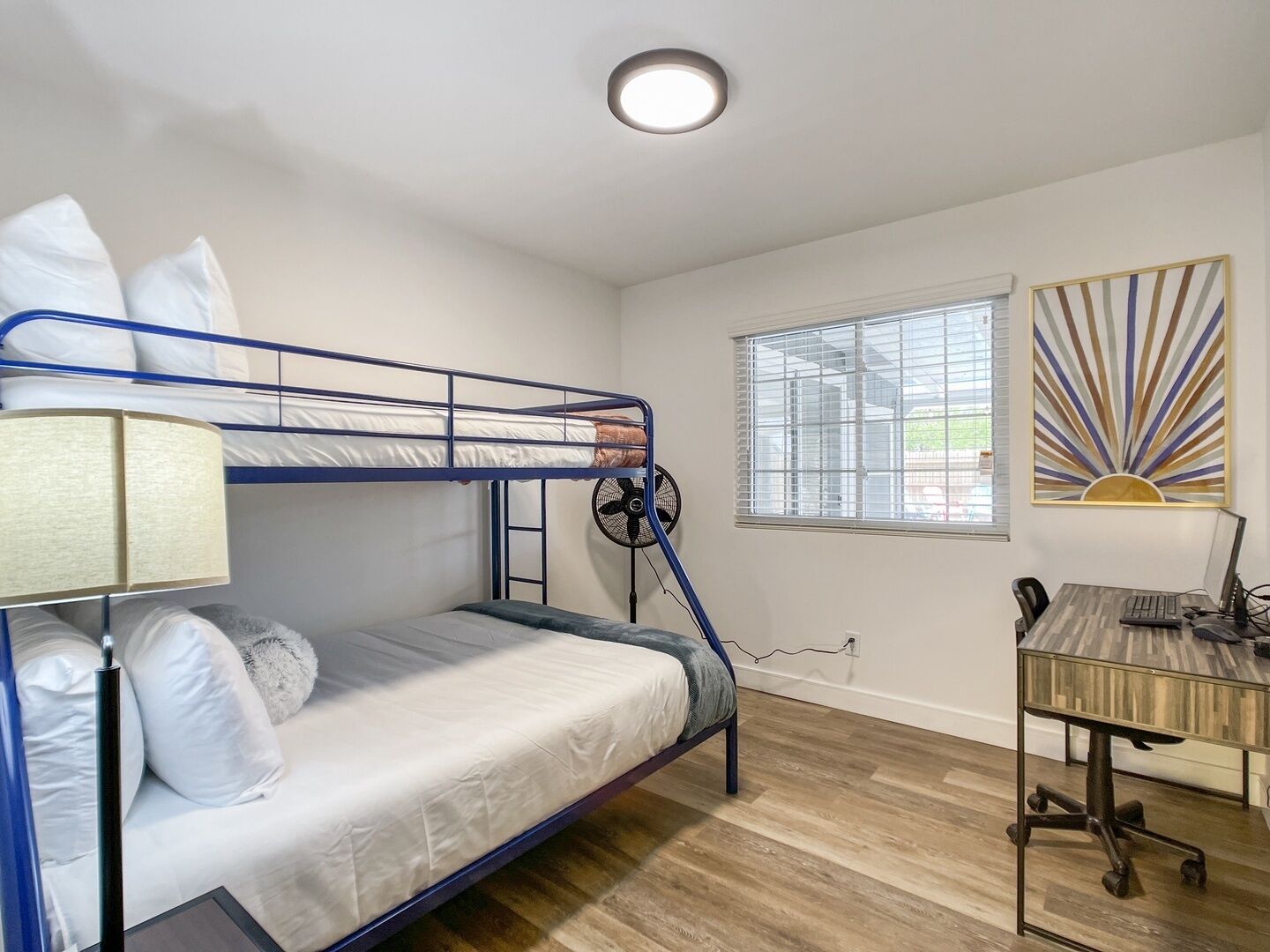 A bunk bed room with a desk and chair, a window with blinds, and a ceiling light.