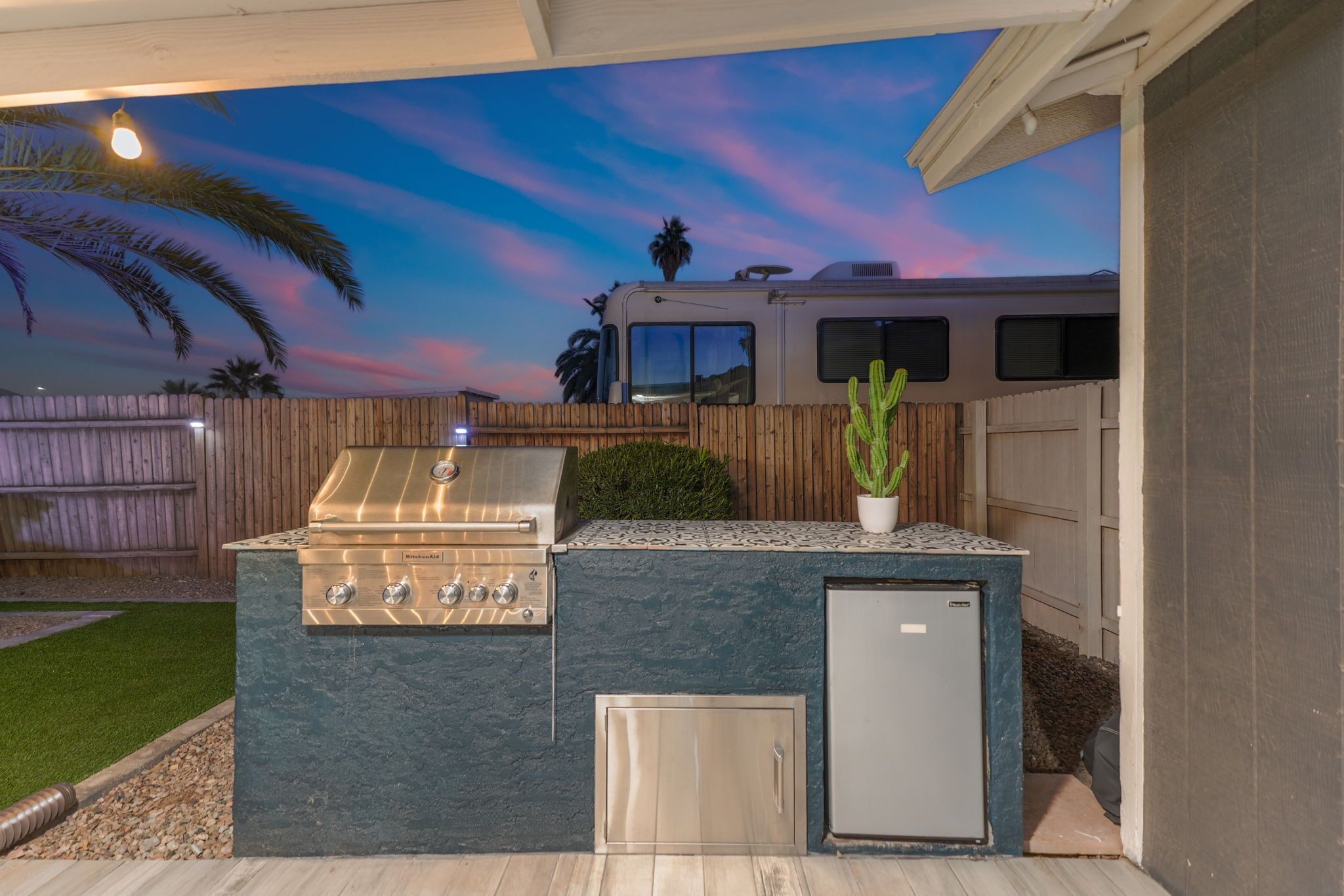 An outdoor kitchen with a grill, a refrigerator, and a wooden fence.