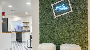 Une cuisine moderne avec un sol à motifs noir et blanc, un mur vert orné d’un néon affichant le message « bonne ambiance », et deux tabourets blancs à l’aspect duveteux, dotés de pieds dorés.